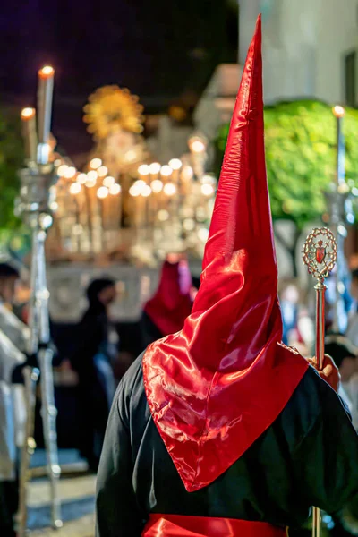 Sivri uçlu kırmızı saten bir başlık ve siyah bir tunik giyen ve elinde süslü gümüş bir asa tutan bir katılımcının arka görüntüsü..