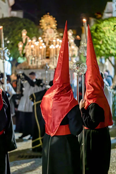 Geleneksel siyah cüppeli ve uzun kırmızı başlıklı bir grup, İspanya 'da dar bir şehir caddesinde, dini bir kutsal hafta geçidinde yürüyorlar..