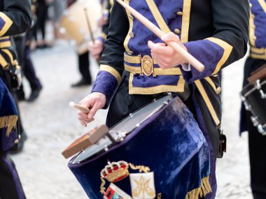 İspanya 'da geleneksel bir dini tören sırasında, mor ve altın rengi tören üniformalı bir müzisyenin ahşap sopalarla trampet çalmasının detaylı görüntüsü...