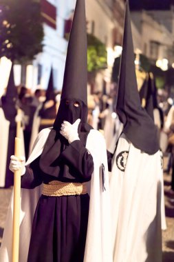 Arcos de la Frontera, Cadiz, İspanya - 17 Nisan 2025: Bir grup Nasıralı siyah başlıklı ve beyaz pelerinli kalabalık bir sokakta yürüyorlar İspanya 'daki kutsal bir hafta dini geçit töreni sırasında ellerinde uzun mumlarla.