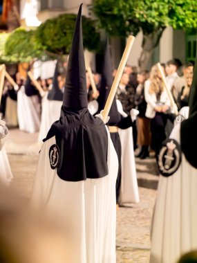 Arcos de la Frontera, Cadiz, İspanya - 17 Nisan 2025: Bir grup Nasıralı siyah başlıklı ve beyaz pelerinli kalabalık bir sokakta yürüyorlar İspanya 'daki kutsal bir hafta dini geçit töreni sırasında ellerinde uzun mumlarla.