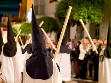 Arcos de la Frontera, Cadiz, İspanya - 17 Nisan 2025: Bir grup Nasıralı siyah başlıklı ve beyaz pelerinli kalabalık bir sokakta yürüyorlar İspanya 'daki kutsal bir hafta dini geçit töreni sırasında ellerinde uzun mumlarla.