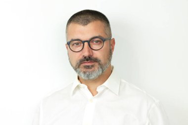 Gözlüklü ve beyaz gömlekli yakışıklı sakallı adam kameraya bakıyor.
