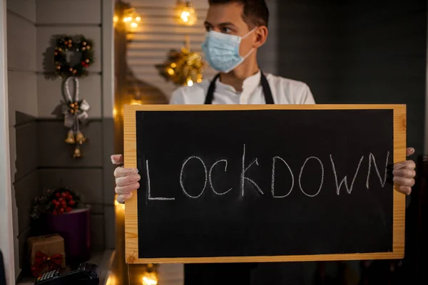 European man waiter in protective mask taking in hands nameplate Lockdown. He depressed and worried about losing his job