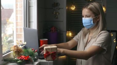 Koruyucu maskeli mutlu bir kadın, elinde hediyeyle karantina sırasında Noel hediyesi almak için evde online alışveriş yapıyor..