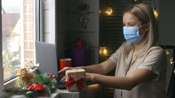 Koruyucu maskeli mutlu bir kadın, elinde hediyeyle karantina sırasında Noel hediyesi almak için evde online alışveriş yapıyor.. — Stok video