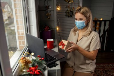 Happy woman in protective mask, taking gift in hand and making  video calling to congratulate Merry Christmas her relatives.  clipart