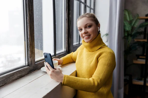 Evde kahve içerken cep telefonu ve kulaklık kullanan genç bir kadın. — Stok fotoğraf