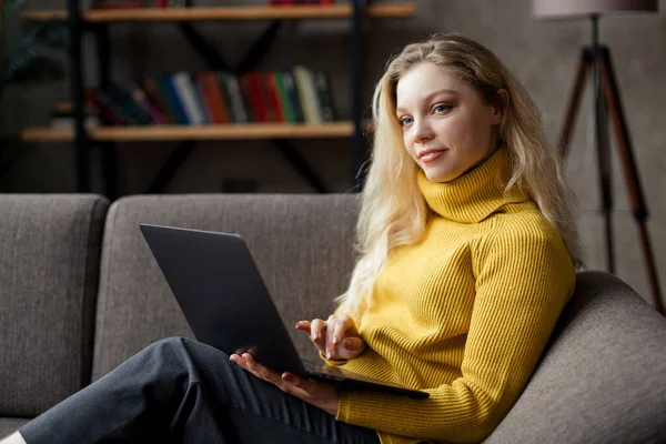 Красива молода жінка працює з дому на ноутбуці, сидячи на дивані у вітальні — стокове фото