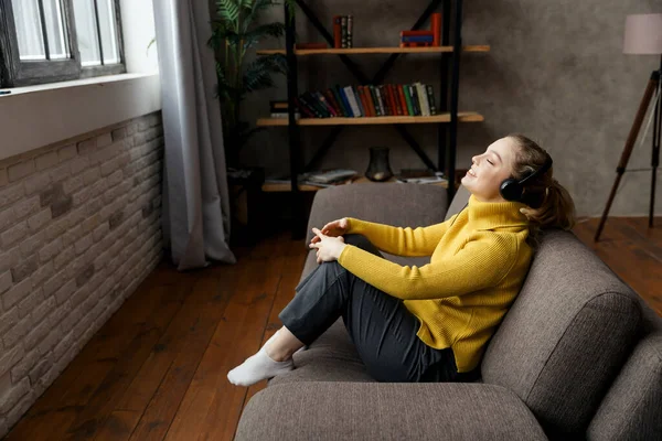 Красива задоволена жінка слухає музику в навушниках, сидячи на дивані вдома — стокове фото