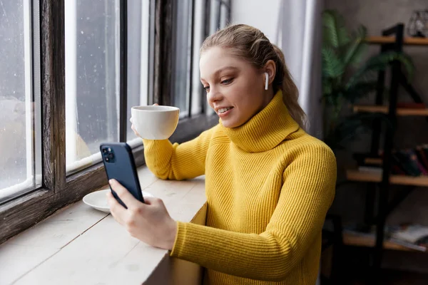 Evde kahve içerken cep telefonu ve kulaklık kullanan genç bir kadın. — Stok fotoğraf