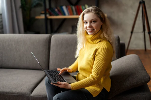 Hermosa joven mujer que trabaja desde casa en un ordenador portátil sentado en el sofá en la sala de estar — Foto de Stock