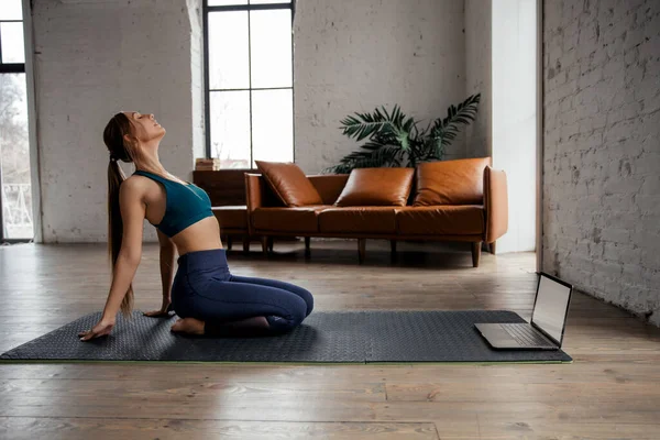 Jovem mulher esportiva em sportswear fazendo ioga no tapete de exercício em casa usando laptop assistindo tutorial. — Fotografia de Stock