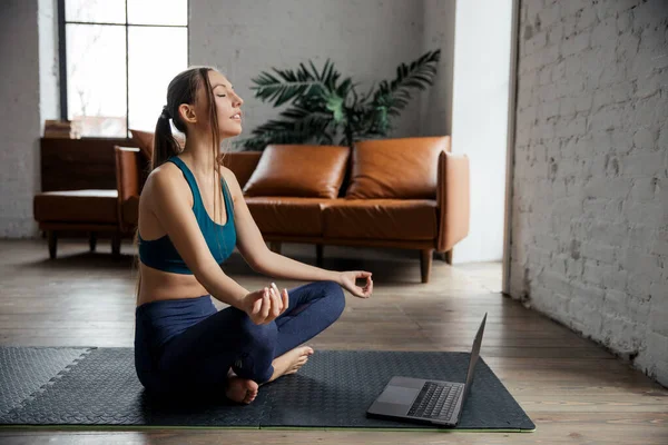 Спортивний костюм струнка жінка-тренер практикує відео онлайн-тренування Хатха-йоги-інструктор в ноутбуці, медитує позу Сухасана, розслабляє дихання . — стокове фото
