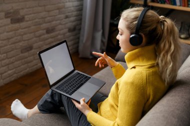 Genç kız oturma odasında çalışıyor, koltukta oturuyor ve kulaklık ve dizüstü bilgisayar kullanıyor..