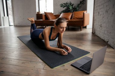 Spor üniformalı genç bir kadın evde kalas egzersizi yapıyor, dizüstü bilgisayarda videolar izliyor ve talimatları tekrarlıyor..