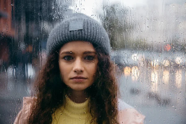Sad young curly woman stay alone at glass with drops in rainy day