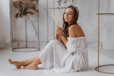 Güzel bir kız, beyaz bir boho elbiseyle yerde oturuyor, tanrıçaya benziyor.