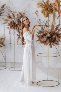 Beyaz boho elbiseli güzel bir kız çiçekliğin yanında duruyor ve etrafına bakıyor.