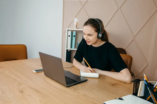 Бізнес-леді в навушниках сидить за столом з ноутбуком, робить нотатки, дивиться вебінар онлайн за відеодзвінком — стокове фото