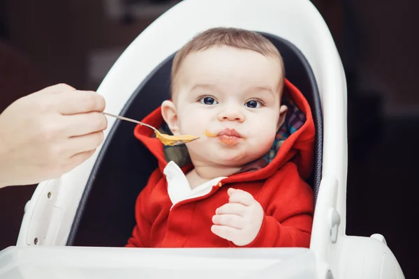Närbild porträtt av söta bedårande kaukasiska lilla baby pojke med mörka svarta ögon sitter i barnstol i köket tittar i kameran äta måltid puré, mamma matar sin son — Stockfoto