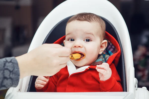 Foto close-up dari anak laki-laki Kaukasia imut dengan mata hitam gelap duduk di kursi tinggi di dapur mencari di kamera makan puree, ibu memberi makan anaknya — Stok Foto