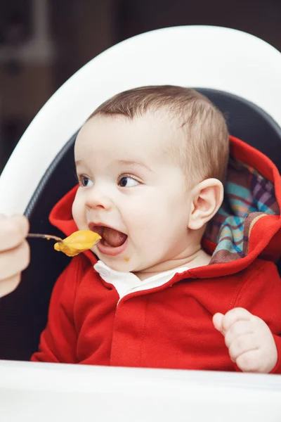 Close-up portrait of cute adorable Caucasian little baby boy with dark black eyes sitting in high chair in kitchen looking away, eating meal puree, mother feeds her son — Stock Image Close-up portrait of cute adorable Caucasian little baby boy with dark black eyes sitting in high chair in kitchen looking away, eating meal puree, mother feeds her son Stock Photo