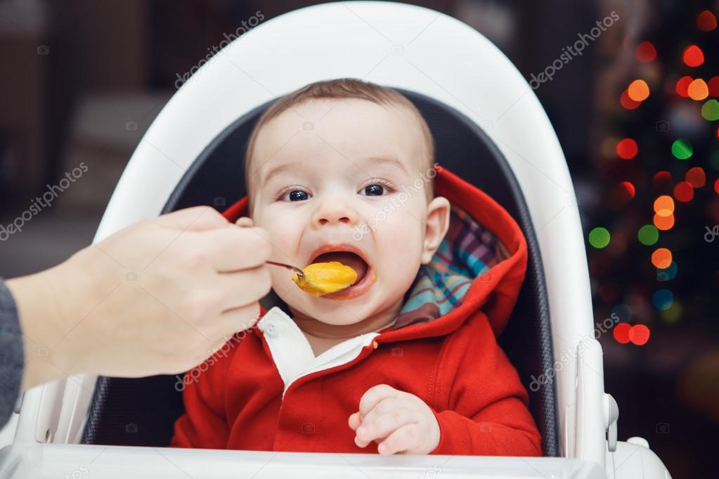 Close-up portrait of cute adorable Caucasian little baby boy with dark black eyes sitting in high chair in kitchen looking in camera eating meal puree, mother feeds her son