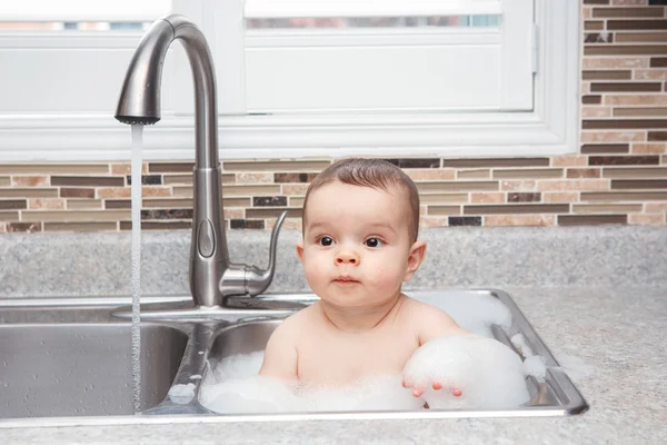 Portret van schattige Kaukasische grappige baby meisje jongen met donkere zwarte ogen zitten in de grote keuken spoelbak met water en schuim in de buurt van venster op weg, zoek levensstijl dagelijks concept — Stockfoto