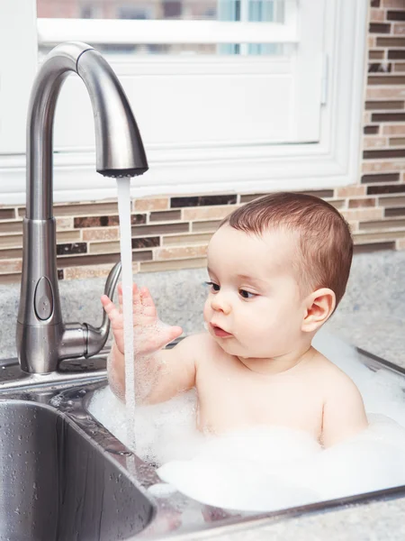 Portret van schattige Kaukasische grappige baby meisje jongen met donkere zwarte ogen zitten in de grote keuken spoelbak met water en schuim in de buurt van venster op weg, zoek levensstijl dagelijks concept — Stockfoto