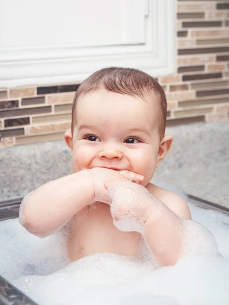 Portret van schattige Kaukasische grappige baby meisje jongen met donkere zwarte ogen zitten in de grote keuken spoelbak met water en schuim in de buurt van venster op weg, zoek levensstijl dagelijks concept — Stockfoto