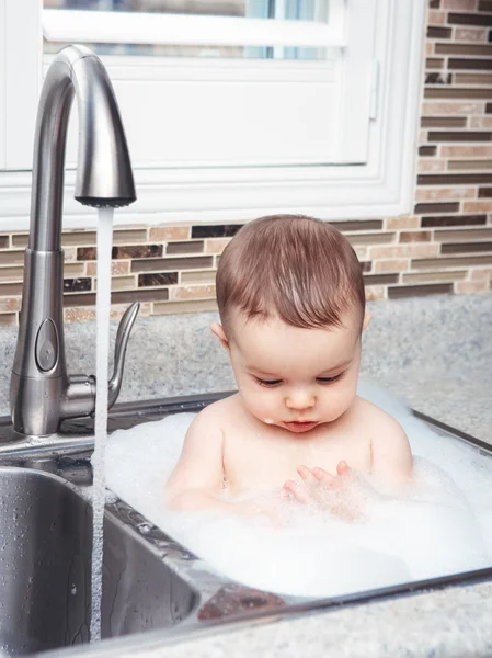 Portret van schattige Kaukasische grappige baby meisje jongen met donkere zwarte ogen zitten in de grote keuken spoelbak met water en schuim in de buurt van venster op weg, zoek levensstijl dagelijks concept — Stockfoto