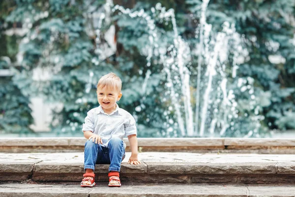 Retrato de lindo adorable divertido niño caucásico niño pequeño en camisa blanca y pantalones vaqueros azules jugando riendo sonriendo divertirse por la fuente en verano al aire libre — Foto de Stock