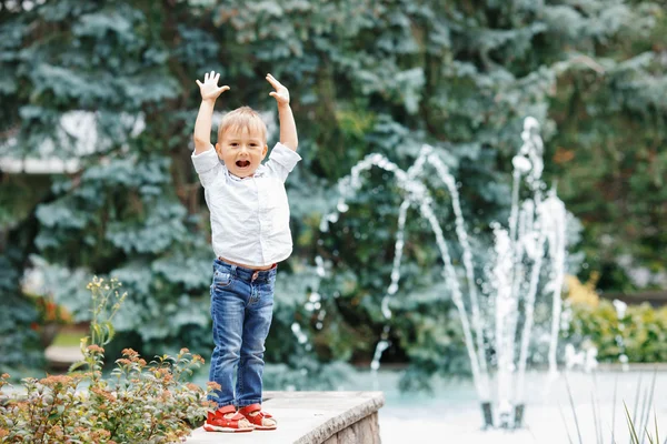 Портрет милого очаровательного смешного белого маленького мальчика в белой рубашке и синих джинсах, играющего смеясь, улыбаясь, веселясь у фонтана летом снаружи — стоковое фото