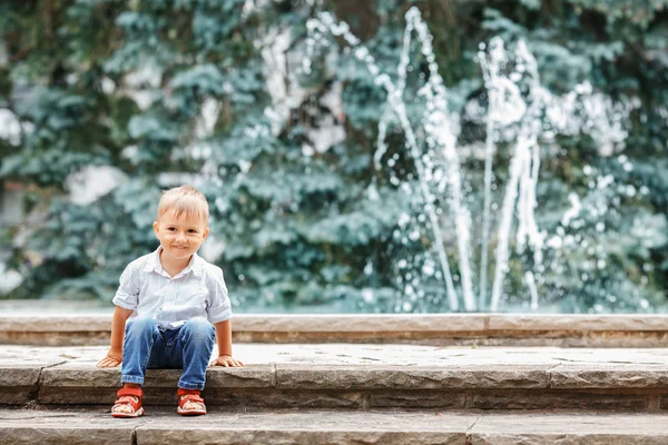 Портрет милого очаровательного смешного белого маленького мальчика в белой рубашке и синих джинсах, играющего смеясь, улыбаясь, веселясь у фонтана летом снаружи — стоковое фото