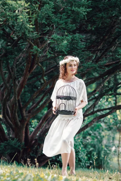 Portrait of beautiful smiling Caucasian young women girl in white long dress with flower chaplet holding empty birdcage in enchanted forest alone, dreamy romantic gothic style — Stock Photo, Image