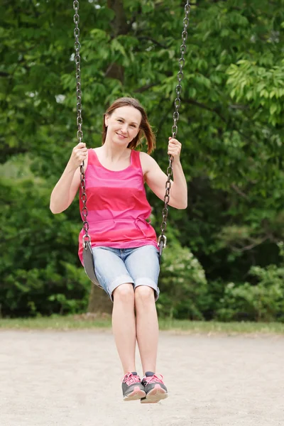 Porträtt av Happy leende ung medelålders kvinna flicka i röd tshirt och jeansshorts på Swing på Backyard Playground utanför på sommardag, livsstil — Stockfoto