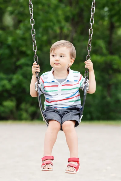 Портрет милого маленького мальчика-малыша, смотрящего в сторону, одетого в футболку и джинсовые шорты на качелях на детской площадке во дворе в летний день, счастливого детского образа жизни — стоковое фото