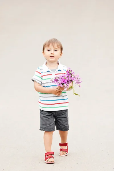 Retrato de um bonito adorável engraçado pequeno menino sorridente criança andando no parque com flores lilás roxas rosa nas mãos no dia de verão brilhante, conceito de feriado dia das mães — Fotografia de Stock