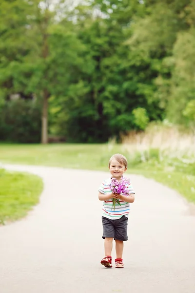 Retrato de un lindo adorable divertido niño sonriente pequeño caminando en el parque con flores de color rosa púrpura lila en las manos en el día de verano brillante, concepto de día festivo de las madres —  Fotos de Stock