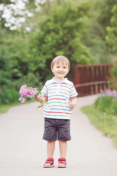 Retrato de um bonito adorável engraçado pequeno menino sorridente criança andando no parque com flores lilás roxas rosa nas mãos no dia de verão brilhante, conceito de feriado dia das mães — Fotografia de Stock