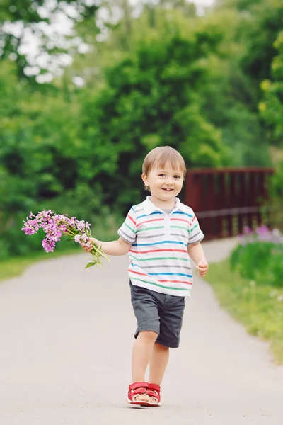 Retrato de um bonito adorável engraçado pequeno menino sorridente criança andando no parque com flores lilás roxas rosa nas mãos no dia de verão brilhante, conceito de feriado dia das mães — Fotografia de Stock