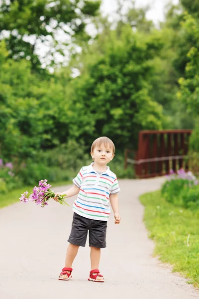 Portrait of a cute adorable funny little smiling boy toddler walking in park with lilac purple pink flowers in hands on bright summer day, mothers day holiday concept — Stock Photo, Image