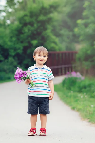 Retrato de un lindo adorable niño sonriente divertido pequeño caminando en el parque con flores de color rosa púrpura lila en las manos en el día de verano brillante, destello de la lente desde arriba, día de las madres —  Fotos de Stock