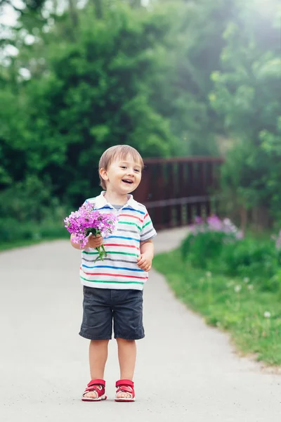 一个可爱的可爱的有趣的小男孩微笑男孩蹒跚学步走在公园与丁香紫色粉红色的花朵在明亮的夏日手，镜头耀斑从上面，母亲节的肖像 — 图库照片