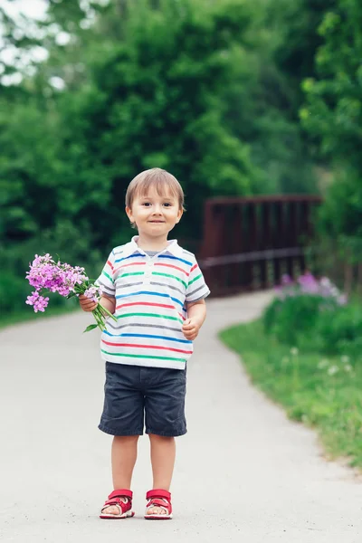 Портрет милого очаровательного смешного маленького улыбающегося мальчика, прогуливающегося в парке с лилово-фиолетовыми розовыми цветами в руках в яркий летний день, День матери концепции праздника — стоковое фото