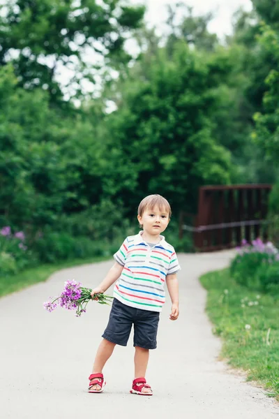 Retrato de um bonito adorável engraçado pequeno menino sorridente criança andando no parque com flores lilás roxas rosa nas mãos no dia de verão brilhante, conceito de feriado dia das mães — Fotografia de Stock