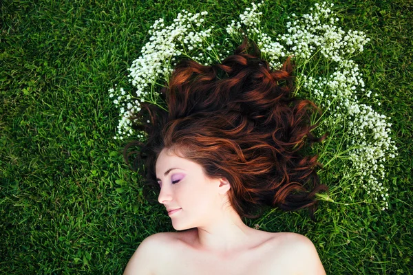 Closeup portrait of young beautiful girl woman with red brown hair lying on grass with white small flowers around her head. View from above top overhead. Concept of spring summer youth happiness — Stock Photo, Image