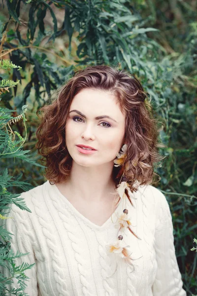 Portrait of beautiful smiling white Caucasian girl woman with long curly hair hazel eyes, in white sweater, feather earring hipster hippie style, among green tree leaves, toned with instagram vsco filters — Stock Photo, Image