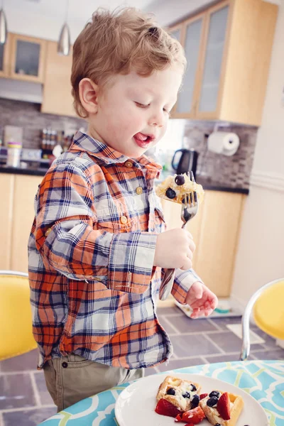 Potret seorang anak yang bahagia makan wafel sarapan dengan buah-buahan dengan garpu di dapur yang cerah di pagi hari menunjukkan lidahnya — Stok Foto